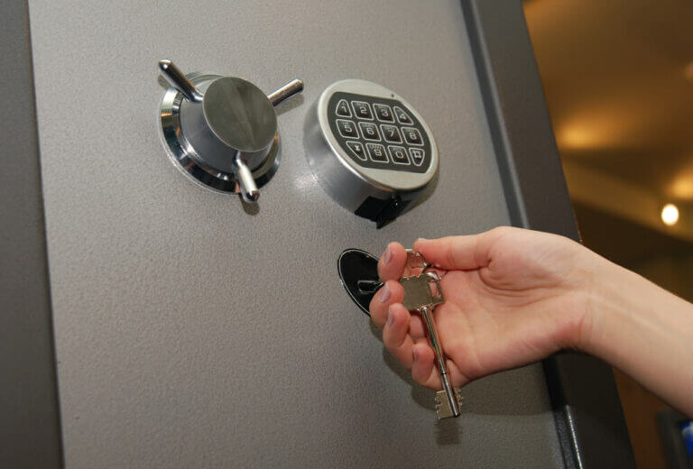 A locksmith's hand is seen inserting a key into a keyhole on a secure safe door. The safe also features a combination lock and a round handle. The background is blurred, focusing attention on the locksmith's hand and the safe components.