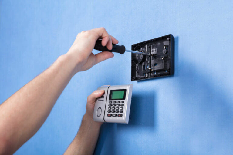 A locksmith is using a screwdriver to install or repair a wall-mounted electronic keypad device against a blue wall. The device features a small screen and a number pad.