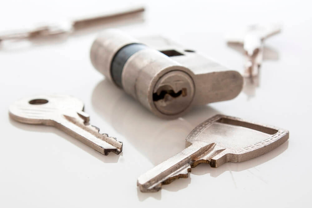 A close-up image shows three keys and a cylindrical lock mechanism arranged on a white surface. Two keys stand upright near the lock cylinder, while the third lies flat in the foreground. Parts of other keys, hinting at locksmith tools, are partially visible in the background.
